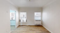 This single-floor unit offers a clean, modern aesthetic defined by warm hardwood flooring and a crisp white color palette that runs throughout the space. The open layout, visible in the dollhouse view, connects the main living areas with a natural flow, providing a sense of openness and versatility.

The living area features multiple windows dressed with white horizontal blinds, allowing generous natural light to fill the room. A wall-mounted HVAC unit ensures year-round comfort, while a contemporary pendant light fixture adds a subtle design accent overhead. A mirrored sliding closet door further enhances the sense of space and light.

The kitchen is well-appointed with gray shaker-style cabinetry, white countertops, a deep stainless steel single-basin sink with a modern pull-down faucet, and a gas range with an overhead ventilation hood. The cabinetry offers ample upper and lower storage, making the kitchen both functional and stylish.

The bedroom is bright and airy, with multiple windows providing cross-ventilation and natural light, complemented by the same warm hardwood flooring found throughout.

The bathroom is a standout feature, showcasing floor-to-ceiling dark marble-effect tile surrounding a full soaking tub, paired with a frosted privacy window. The contrast of rich dark tile against the white fixtures and hardwood flooring creates a striking, spa-like atmosphere.
