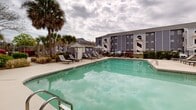 This contemporary residential complex offers a blend of comfort and leisure. The centerpiece is a spacious, resort-style swimming pool with a generous deck area, perfect for sunbathing and relaxation. The pool is surrounded by comfortable lounge chairs and umbrellas, providing ample seating options for residents.

The architecture is characterized by multi-story buildings with a mix of gray and white exterior finishes, creating a clean and modern aesthetic. Large windows and balconies offer residents outdoor spaces and natural light. Distinctive arched windows on the upper floors add architectural interest.

Landscaping plays a significant role in the complex's ambiance, with mature palm trees dotting the grounds, lending a tropical feel to the environment. Well-maintained shrubbery and green spaces further enhance the property's appeal.

For outdoor entertaining, the complex features communal seating areas with modern patio furniture, ideal for socializing or enjoying a meal al fresco. A perimeter fence ensures privacy and security for residents.

The property appears to offer additional amenities, possibly including a clubhouse or communal building, as seen in the images. Overall, this apartment complex presents an attractive living space with a focus on outdoor enjoyment and community living.