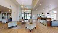 Located at 7990 E Snyder Rd in Tucson, Arizona, this single-story community clubhouse offers a sophisticated and welcoming environment designed for both relaxation and social gathering. The main lounge area commands attention with its soaring ceilings and a dramatic coffered ceiling detail, anchored by a cluster of sculptural globe pendant lights that cast a warm, inviting glow. Light wood-tone flooring runs throughout, enhancing the bright and airy atmosphere created by an abundance of natural light.

Expansive floor-to-ceiling black-framed glass walls and French doors dominate one side of the lounge, offering direct views of the outdoor pool area and seamlessly connecting the interior to the exterior. The seating arrangement features contemporary tufted armchairs and a stylish circular side table atop a patterned area rug, creating an elegant yet comfortable social space.

Opposite the lounge, a dedicated reception and service counter area provides a functional hub, accented by decorative wall texturing. A fully equipped kitchenette adjoins the main space, outfitted with warm wood cabinetry, white stone countertops, open floating shelves, and black-framed windows overlooking lush palm trees. The kitchenette includes a sink, microwave, and coffee station, making the space well-suited for community events and everyday use.