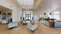 Located at 7990 E Snyder Rd in Tucson, Arizona, this single-story community clubhouse offers a sophisticated and welcoming environment designed for both relaxation and social gathering. The main lounge area commands attention with its soaring ceilings and a dramatic coffered ceiling detail, anchored by a cluster of sculptural globe pendant lights that cast a warm, inviting glow. Light wood-tone flooring runs throughout, enhancing the bright and airy atmosphere created by an abundance of natural light.

Expansive floor-to-ceiling black-framed glass walls and French doors dominate one side of the lounge, offering direct views of the outdoor pool area and seamlessly connecting the interior to the exterior. The seating arrangement features contemporary tufted armchairs and a stylish circular side table atop a patterned area rug, creating an elegant yet comfortable social space.

Opposite the lounge, a dedicated reception and service counter area provides a functional hub, accented by decorative wall texturing. A fully equipped kitchenette adjoins the main space, outfitted with warm wood cabinetry, white stone countertops, open floating shelves, and black-framed windows overlooking lush palm trees. The kitchenette includes a sink, microwave, and coffee station, making the space well-suited for community events and everyday use.