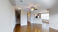 This two-story residence offers a thoughtfully designed open-concept main floor that combines comfort and functionality. Rich hardwood flooring extends throughout the living area, creating a warm and inviting atmosphere. A ceiling fan with a built-in light fixture anchors the living space, providing both airflow and ambient lighting. The staircase, positioned near the entryway, features carpeted treads and leads to the upper floor, offering a clear separation between living and private spaces. The kitchen is centrally positioned and features a peninsula-style counter that serves as both a workspace and a casual dining area. Pendant lighting hangs above the peninsula, adding a refined touch to the space. The kitchen is equipped with stainless steel appliances, including a refrigerator and microwave, and is framed by light-toned cabinetry. A large window in the adjoining area floods the space with natural light, enhancing the open and airy feel of the main floor. Neutral gray walls throughout provide a clean, versatile backdrop that complements a variety of interior styles. White trim and baseboards add a crisp finishing detail, contributing to the overall polished aesthetic of this well-appointed home.