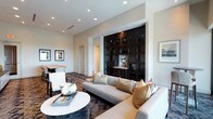 This single-floor space is thoughtfully designed to blend comfort and elegance in a contemporary lounge setting. The open layout seamlessly connects a relaxed seating area with a casual dining nook, creating a versatile environment suited for both socializing and quiet relaxation.

The living area centers around a generously sized sectional sofa in neutral gray upholstery, accented with an assortment of warm-toned throw pillows. A round white coffee table serves as a clean focal point, styled with decorative books and fresh floral accents. Underfoot, a richly patterned area rug in deep earth tones adds visual depth and warmth.

A dramatic built-in cabinetry unit in dark espresso wood dominates one wall, featuring latticed wine storage, glass-front display shelving, and a marble-topped surface — functioning as both a design statement and a practical bar area.

Adjacent to the lounge, a compact dining space accommodates a live-edge wood table paired with sleek white chairs, ideal for small gatherings. Framed artwork punctuates the neutral walls throughout. Recessed lighting illuminates the space evenly, complementing the clean white ceiling and creating an inviting, polished ambiance.