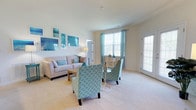 Located at 100 Isabelle Isle in Dover, Delaware, this single-floor residence offers a comfortable and thoughtfully designed living environment. The living area serves as the heart of the space, showcasing plush wall-to-wall carpeting that adds warmth underfoot. Large windows dressed with soft aqua curtains flood the room with abundant natural light, creating an airy and inviting atmosphere throughout the day. A glass-paneled door provides additional brightness and easy access to the outdoors. The living area is generously proportioned, comfortably accommodating a full seating arrangement that includes a neutral-toned sofa and patterned accent chairs in a classic quatrefoil design, lending the space a coastal, relaxed aesthetic. Ocean-inspired wall art in cool blues and greens reinforces the light, serene ambiance. The neutral, light-toned walls and soft color palette contribute to a calm and open feel throughout. The layout is practical and functional, with the single-floor design ensuring easy accessibility and seamless flow between spaces. Overall, this residence at Isabelle Isle offers a bright, tranquil setting ideal for comfortable everyday living in Dover, DE.