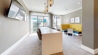 Located at 300 Central Ave in the heart of downtown Sarasota, Florida, this thoughtfully designed single-floor communal space offers a sophisticated blend of functionality and contemporary style. The layout encompasses several distinct zones, each tailored to different needs while maintaining a cohesive aesthetic throughout.

The central gathering area is anchored by a long, sleek white communal table surrounded by modern white chairs, illuminated overhead by a trio of amber globe pendant lights that add warmth to the neutral palette. This space is bathed in natural light streaming through floor-to-ceiling windows, which frame views of the surrounding downtown streetscape. Sheer drapery softens the light and enhances the airy atmosphere.

Adjacent to the main table area, a cozy lounge nook features a bold yellow curved booth paired with a small round table and casual seating, accented by a decorative geometric wall panel. Patterned carpet tiles in warm taupe tones carry throughout the space, complementing the greige walls and white trim.

Private offices enclosed in glass-framed black-trimmed partitions provide focused work areas, one of which features a striking yellow botanical wallcovering. A smaller workspace nook with a built-in desk and window views offers an additional quiet retreat. The space also includes a service counter area and direct access via double doors, making it well-suited for collaborative or professional use.