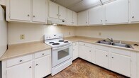 Located at 529 W Grand Ave in Dayton, Ohio, this single-floor unit offers a straightforward, functional layout suited for comfortable everyday living. The floor plan, visible in the 3D overview, reveals a well-organized arrangement of interconnected spaces including a living area, kitchen, and additional rooms accessible via a central corridor.

The living areas are finished with wall-to-wall beige carpeting and neutral cream-colored walls, creating a calm, understated ambiance throughout. One of the carpeted rooms features a tall, narrow window dressed with vertical blinds, allowing natural light to filter in and providing a view of the surrounding exterior. The ceiling features a paneled design with subtle beam detailing visible in several areas.

The kitchen is equipped with white cabinetry offering ample upper and lower storage, light-toned laminate countertops in an L-shaped configuration, a white electric coil range with an overhead exhaust hood, and a stainless steel sink. The kitchen flooring consists of durable vinyl tile in a warm, mottled pattern.

An entry corridor connects the main spaces, featuring a front door with an adjacent window panel that brings in additional light. A closet is also accessible near the living area. HVAC vents are present throughout, indicating central climate control. Overall, the space maintains a clean, neutral palette ready for personalization.