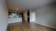 This single-floor space showcases an open-concept layout that seamlessly connects the living area and kitchen. The kitchen is outfitted with dark, espresso-toned cabinetry that extends to the upper walls, stainless steel appliances including a refrigerator, stove, and built-in microwave, and a granite-topped island with a breakfast bar overhang. A stone tile backsplash adds texture and visual interest behind the range. Pendant lighting hangs above the island, complemented by a track lighting fixture, providing focused illumination over the workspace.

The living area extends generously from the kitchen, finished with warm-toned hardwood flooring that runs continuously throughout the space, contributing to a cohesive and inviting atmosphere. Neutral gray walls provide a versatile backdrop. A built-in shelving nook adds functional storage near an interior doorway. A secondary door, finished in white with classic panel detailing, leads to an adjacent room. The overall ambiance is modern yet understated, with a balanced combination of dark and light elements creating a polished, contemporary feel.