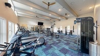 Located at 13611 Perry Rd in Houston, TX 77070, this single-story fitness center is designed to support a full range of exercise routines in a bright, open environment. The space features notably high ceilings with exposed decorative beams, recessed lighting, and ceiling fans that contribute to an airy, comfortable atmosphere during workouts.

Large windows line multiple walls, including an expansive bank of floor-to-ceiling windows that flood the space with natural light and provide outdoor views of the surrounding landscape. Additional clerestory windows near the roofline further enhance brightness throughout the facility.

The flooring consists of a patterned carpet tile in deep blue, teal, and purple tones, adding a modern aesthetic to the interior. Equipment is well-distributed across the open floor plan and includes treadmills, elliptical trainers, a stationary cycling bike, cable machines, a functional trainer with suspension straps, strength machines, and free weight stations. Wall-mounted mirrors expand the visual scale of the space and support proper form during training.

A dedicated storage area with cubbies and a counter provides space for towels and accessories. A wall-mounted television and built-in speakers complete the facility, ensuring an engaging and fully equipped workout environment.