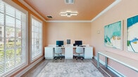 Located at 10407 US-31 in Spanish Fort, Alabama, this single-room office space offers a thoughtfully designed and fully equipped work environment. The room is finished in soft peach and blush tones with a warm-toned ceiling and crisp white crown molding, creating an inviting yet professional ambiance throughout.

The primary workstation area features a long, white built-in desk with cabinet storage beneath, accommodating two computer monitors and ergonomic rolling chairs — ideal for collaborative or side-by-side work. A sleek modern flush-mount light fixture illuminates the workspace from above, complementing the natural light that pours in through large windows fitted with white horizontal blinds.

A seating nook near the glass-paneled entry door provides a comfortable lounge area with two light blue upholstered armchairs positioned on a soft, neutral area rug that anchors the space. This area is equally well-lit, benefiting from both the entry door's window and an additional side window with outdoor views of surrounding greenery.

The walls are adorned with contemporary abstract artwork in cool aqua and teal tones, offering an artistic contrast to the warm wall color. Light wood-style flooring runs throughout, tying the space together cohesively. The office is well-ventilated with ceiling vents and is equipped with a fire safety system, ensuring both comfort and compliance.