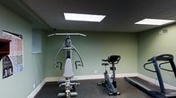 This single-floor fitness space is purpose-built for exercise and personal wellness. The room is finished with sage green walls and white baseboards that create a calm, focused atmosphere conducive to working out. Overhead, flush-mounted panel lighting provides bright, even illumination throughout the space, ensuring excellent visibility during any exercise routine.

The room is equipped with a multi-function cable and weight resistance training machine, offering a wide range of strength exercises from a single unit. An upright stationary exercise bike is positioned nearby, ideal for low-impact cardiovascular workouts. A treadmill along the far wall rounds out the cardio offerings, providing options for walking or running. The flooring is covered with dark rubber or foam fitness matting, offering both cushioning and slip resistance — a practical and safety-conscious choice for a workout environment.

The space features a clean, uncluttered layout that allows for comfortable movement between equipment. The combination of strength and cardio machines makes this a well-rounded fitness area suited for a complete workout routine. The overall design prioritizes function and practicality, making it an efficient and fully equipped space for regular physical activity.