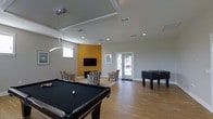 This single-floor recreational space is thoughtfully designed to serve as a lively hub for social interaction and entertainment. The room features light hardwood flooring that runs throughout, complemented by neutral gray walls and a bold golden-yellow accent wall that adds a pop of color and visual interest. Recessed lighting and a sleek pendant fixture illuminate the space evenly, enhancing the open, airy feel created by the high tray ceiling.

At the center of the room, a full-size pool table commands attention, while a foosball table nearby offers additional gaming entertainment. A comfortable lounge area is anchored by a pair of colorful patterned armchairs positioned in front of the wall-mounted flat-screen television, creating an inviting spot for relaxation. Natural light pours in through multiple windows and a set of glass French doors, which open to an exterior area and contribute to the room's bright, welcoming ambiance.

The layout is spacious and well-organized, allowing for easy movement between activity zones. The combination of game tables, comfortable seating, entertainment fixtures, and abundant natural light makes this a versatile, engaging space well-suited for community gatherings and recreational use.
