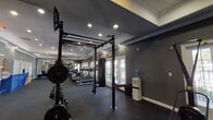 This single-story fitness center is designed to accommodate a comprehensive range of workout needs within a spacious, open layout. The interior is finished with dark charcoal-toned walls, crown molding, tray ceilings, and recessed lighting, creating a focused yet polished athletic environment. Durable rubber flooring spans the entire space, providing both safety and functionality.

The cardio zone lines one wall and features multiple treadmills and elliptical machines equipped with individual digital displays. Large French doors and windows along this wall allow abundant natural light to filter in while offering outdoor views. Full-length mirrors on the opposite wall visually expand the space and allow users to monitor their form.

The strength training area includes a multi-functional power rack with a pull-up bar, a barbell with weight plates, a heavy punching bag, a cable crossover machine, and an adjustable bench. Ceiling fans provide supplemental air circulation throughout.

Adjacent to the fitness floor is an entry corridor featuring a modern automated package locker system in a blue-and-white finish, with glass partitions opening toward a bright communal lounge area beyond. A water cooler station is also available within the fitness space for user convenience.