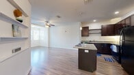 Located at 130 Town Center Blvd in Clermont, FL, this one-bedroom, one-bathroom residence offers a thoughtfully designed single-floor layout with modern finishes throughout. The open-concept kitchen and living area serves as the heart of the home, featuring rich dark-espresso cabinetry, granite countertops, a center island, pendant lighting, and black appliances. Track lighting and recessed fixtures illuminate the kitchen, while the living area benefits from a ceiling fan and natural light streaming through a front-facing window. Wood-style plank flooring extends throughout the main living spaces, adding warmth and continuity. Built-in wall shelving in the living area provides functional display and storage space. A hallway connects the main living area to the private quarters, with access to the bedroom, bathroom, closet, and in-unit laundry room. The bedroom features plush carpeting, a ceiling fan with light, and a window offering natural light. The bathroom is equipped with granite-topped vanity cabinetry and a tub-shower combination. The dedicated laundry room includes a full-size washer and dryer. Clean white walls and consistent finishes throughout create a bright, cohesive living environment.
