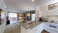 This single-floor residence features two bedrooms and two bathrooms within a thoughtfully designed open-concept layout. The kitchen serves as a central hub of the home, boasting white cabinetry, granite-style countertops, and a wraparound breakfast bar that seamlessly connects to the living area. Under-cabinet appliances, including a built-in microwave, add functional convenience to the space. A decorative pendant light anchors the kitchen, providing warm, focused illumination over the counter.

The open flow between the kitchen and living room creates a spacious, cohesive feel throughout the main living area. The living room is carpeted and bathed in natural light from large windows dressed with grey curtains, while a sliding glass door leads to a private balcony, offering outdoor access and additional light. Accent walls in a deep grey tone add visual interest and depth to the space.

Crown molding details and a textured ceiling contribute subtle architectural character throughout. The overall ambiance is bright, welcoming, and functional, making efficient use of the available floor space across a single level.