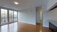 This single-floor space offers a clean, modern aesthetic defined by warm hardwood flooring that extends throughout the open living area. Large floor-to-ceiling windows along one wall flood the space with natural light and provide direct access to a private balcony, offering outdoor views and a strong sense of connection to the surrounding environment. The neutral white walls and smooth ceilings create a bright, airy atmosphere that enhances the feeling of openness. A flush-mount ceiling light provides even ambient illumination, complementing the natural light. The layout flows naturally from the main living area toward additional rooms accessible through open doorways, suggesting a well-organized and functional floor plan. Crisp white baseboards and clean transitions between spaces contribute to the polished, contemporary look. The overall ambiance is calm and inviting, with a minimalist character that allows for flexible use and personalization of the space.