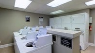 Located at 2979 Green Arbor Ln in Dublin, Ohio, this shared laundry facility offers a clean, well-organized, and fully equipped space designed for efficient use. The room features a row of top-loading washing machines arranged along one side, complemented by a bank of stacked dryers lining the back wall, providing ample laundry capacity for residents. A built-in white folding counter runs along the center of the room, offering a convenient surface for sorting and folding laundry. The space is finished with neutral gray walls and a suspended drop ceiling with recessed fluorescent panel lighting, ensuring the room is consistently bright and well-lit throughout. The flooring is smooth and easy to clean, with caution signage posted to alert users to slippery conditions when wet, reflecting a commitment to safety. Informational notices are displayed on the walls, keeping users informed of facility rules and guidelines. The overall layout is practical and spacious, allowing multiple users to move comfortably through the room simultaneously. This well-maintained laundry facility provides everything needed for a straightforward and convenient laundry experience.