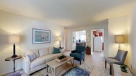 This single-floor residence at Whitehall Gardens offers a thoughtfully designed layout with a warm, coastal-inspired aesthetic throughout. The living room serves as the heart of the home, featuring neutral carpet flooring, soft wall tones, and a sliding glass door framed by blue curtains that invite natural light and provide access to the outdoors. The open sightlines connect the living area to both the kitchen and a secondary room, enhancing the sense of flow and spaciousness.

The kitchen is equipped with rich wood cabinetry, light countertops, a stainless steel sink, a full-size refrigerator, and a dishwasher, with a window above the sink providing additional natural light. The bedroom is generously sized with plush carpeting, a large window dressed in blue drapes, and ample room for furnishings. A second bedroom or flex space functions well as a home office, featuring a window with exterior views and enough room to accommodate a substantial desk and seating area. The bathroom includes a tub-shower combination, a wood vanity with a mirror, and a window for ventilation. Consistent neutral tones and carpeted floors throughout lend the residence a cohesive and comfortable character.