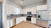 This single-story space offers a clean, contemporary design throughout, highlighted by a cohesive neutral color palette of soft grays, crisp whites, and blue-gray accents. The kitchen serves as a central feature, showcasing white shaker-style cabinetry extending to the ceiling for generous storage, granite countertops with subtle speckled detailing, and a full suite of stainless steel appliances including a French door refrigerator with ice maker, electric range, over-the-range microwave, and dishwasher. A gray tile backsplash and light wood-look flooring complete the polished look. Adjacent to the kitchen, a dedicated laundry area houses a stacked washer and dryer unit behind a blue-gray door, maintaining a tidy and organized flow. The bathroom features a granite-topped vanity with an undermount sink, a large mirror, wood-look tile flooring, and a tub-shower combination with glass enclosure. Bedrooms are carpeted for comfort and receive ample natural light through large windows with views of the surrounding exterior. Throughout the space, blue-gray door and baseboard accents provide a consistent design detail that ties the interior together.