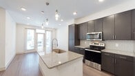 The Malibu floor plan is a thoughtfully designed one-bedroom, one-bathroom unit that maximizes its single-floor layout with an open-concept living and kitchen space. The kitchen serves as a central highlight, anchored by a large island with granite countertops, an integrated sink, and striking glass pendant lights overhead. Dark espresso cabinetry extends to the ceiling, paired with a neutral tile backsplash and a full suite of stainless steel appliances, including a range, microwave, and refrigerator. Wood-style plank flooring runs throughout the main living areas, lending warmth and continuity to the space.

The open layout connects the kitchen seamlessly to the living area, which benefits from recessed lighting and access to a glazed entry door that invites natural light. The bedroom is carpeted for comfort, features a ceiling fan, and includes multiple windows with blinds that provide ample natural light and outdoor views.

The bathroom is finished with wood-style flooring, granite countertops on the vanity with dark cabinetry, and a full soaking tub and shower combination with neutral tile surround. Adjacent to the bathroom, a walk-in closet adds practical storage. The overall aesthetic is clean and contemporary throughout.