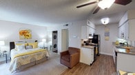 This single-floor studio offers an efficient and thoughtfully designed open-concept layout that seamlessly integrates the living, sleeping, and kitchen areas into one cohesive space. The sleeping area features plush carpeting and is accented by warm, cheerful décor, with a ceiling fan overhead providing both comfort and ambient lighting. A smooth transition leads to the kitchen, which is outfitted with white cabinetry, a black refrigerator, an electric range, and a compact countertop workspace — providing all the essentials for everyday meal preparation. The flooring in the kitchen area transitions to hardwood-style planking, adding a clean visual contrast to the carpeted living zone. A notable convenience of this studio is the in-unit stacked washer and dryer, tucked neatly into a dedicated utility alcove, eliminating the need for shared laundry facilities. A doorway visible from the main living area leads to what appears to be a bathroom or additional storage, keeping the layout functional and organized. The textured ceiling and neutral wall tones create a bright, airy atmosphere throughout, while the open floor plan maximizes the sense of space, making this studio a practical and comfortable single-floor residence.