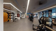 This expansive fitness center is thoughtfully designed to accommodate a full range of workout styles across a single open floor. The space is divided into distinct functional zones, each outfitted with purpose-specific equipment. Linear LED lighting runs along the ceiling throughout, providing bright, even illumination across the entire facility, while ceiling fans contribute to air circulation and comfort.

The main training floor features durable rubber flooring throughout, offering both safety and resilience. A dedicated cardio zone lines the exterior wall, where large floor-to-ceiling windows flood the area with natural light and outdoor views. Bold, full-color motivational murals and graphics cover the surrounding walls, creating an energizing atmosphere. Treadmills, stationary bikes, and stair climbers are positioned to take full advantage of the natural light and views.

A strength training section occupies the central portion of the facility, housing a selection of cable machines and selectorized equipment arranged to maximize usable floor space. A separate functional training area includes a heavy-duty power rack set against a warm wood-paneled accent wall, TRX suspension trainers, a tire for functional drills, and a rowing machine. Additional free weights and accessories are neatly organized throughout, completing a well-rounded training environment.