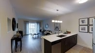Located at 201 Willett Ave in Port Chester, New York, this single-floor residence offers a thoughtfully designed open-concept layout that seamlessly connects the kitchen, dining, and living areas. The kitchen serves as a central hub, anchored by a large island with a white quartz countertop, an undermount sink with a modern pull-down faucet, dark espresso cabinetry, and a stainless steel dishwasher. A sleek multi-pendant chandelier hangs above the island, adding a refined touch to the space. The open living and dining area is finished with smooth hardwood-style flooring that flows throughout, creating a cohesive and contemporary feel. The living area is bathed in natural light, drawn in through a large sliding glass door that opens to a private balcony, framed by floor-length patterned drapes. The neutral color palette of soft grays and whites throughout the space contributes to a clean, modern aesthetic. Recessed lighting complements the natural light, ensuring the space remains bright and welcoming. The overall ambiance is polished and functional, ideal for everyday living and entertaining alike.