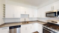 Located at 5300 Westbrook Blvd in East Islip, NY, this well-appointed single-floor residence offers a clean, modern aesthetic throughout. The kitchen is a standout feature, showcasing crisp white shaker cabinetry extending to the ceiling for ample storage, taupe countertops, and a full suite of stainless steel appliances including a gas range, over-the-range microwave, dishwasher, and refrigerator. Rich wood-look flooring runs through the kitchen and into the open living area, which is bright and airy thanks to large double-hung windows that bring in generous natural light and offer views of the surrounding neighborhood. The bedroom is finished with soft wall-to-wall carpeting and features a spacious walk-in closet outfitted with wire shelving and storage racks on multiple walls. The bathroom is cleanly designed with large-format tile flooring, a white vanity with ample cabinet storage, and an oval mirror with contemporary wall sconce lighting. A dedicated laundry room provides added convenience with a full-size side-by-side washer and dryer and wire shelving above for storage. The overall ambiance is fresh and move-in ready, with a neutral color palette and consistent modern finishes throughout.