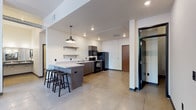 Located at 520 4th Street in Cheney, Washington, this single-floor unit presents a practical and modern industrial aesthetic throughout its layout. The open-concept kitchen and living area serves as the central hub of the space, featuring large multi-pane windows that bring in natural light and views of surrounding greenery. The kitchen is well-equipped with dark cabinetry in a taupe-gray finish, black appliances including a coil-top range, over-range microwave, dishwasher, and full-size refrigerator, along with a double-basin sink and light-toned countertops. Open metal wall shelving adds a utilitarian touch. A peninsula with bar-height seating connects the kitchen to the living area, which accommodates seating and benefits from the same generous window presence. Polished concrete-style flooring runs throughout the main living areas, lending an urban feel to the space. Two bedrooms feature carpeted floors, built-in storage bed frames, and windows with outdoor views. Two bathrooms each include a tub-shower combination with tiled surrounds and clean white fixtures. A glass-paneled door adds a contemporary architectural detail near the entry.