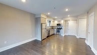 This well-appointed 1-bedroom, 1-bathroom residence is thoughtfully designed across a single floor, offering a seamless blend of modern aesthetics and functional living. The open-concept layout connects the living area and kitchen, creating a spacious and airy atmosphere enhanced by recessed lighting throughout.

The kitchen is a standout feature, outfitted with a full suite of stainless steel appliances, including a refrigerator, range with oven, built-in microwave, and dishwasher. Light wood-toned cabinetry provides ample storage, while the clean countertops and neutral color palette contribute to a crisp, contemporary look. A window above the sink allows natural light to brighten the workspace.

Rich, dark hardwood-style flooring runs throughout the main living area, offering a warm contrast to the soft grey walls and white trim. The open living space provides generous room for furnishing and personalization. Recessed ceiling lights ensure consistent, even illumination across the entire area.

A hallway visible from the main living space leads to additional rooms, maintaining a well-organized and private layout. The overall ambiance is clean, modern, and move-in ready, making this residence an ideal space for those seeking contemporary comfort and efficient design.