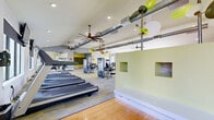 Located at 6465 142nd Avenue North in Clearwater, Florida, this well-appointed fitness center offers a generously sized, single-floor layout designed to accommodate a full range of workout activities. The space is defined by its open, airy feel, with vaulted ceilings, exposed industrial-style ductwork, large ceiling fans, and recessed lighting that collectively create a bright and energetic atmosphere. Stylish copper pendant light fixtures add a distinctive design touch throughout.

The flooring transitions between warm hardwood and patterned carpet tiles in yellow and gray, visually delineating different workout zones. The cardio area is equipped with a row of treadmills and multiple elliptical cross-trainers, all positioned near large windows that allow natural light and outdoor views. A dedicated cycling zone features spin bikes alongside a wall of built-in shelving and bench seating. Strength training is well-represented with a selection of selectorized machines and a free weight rack with a wall mirror.

A functional training area at one end of the facility includes a punching bag, TRX suspension trainers, a medicine ball, and open floor space on hardwood, ideal for dynamic exercises. Wall-mounted televisions are positioned throughout the space. The interior is further enlivened by playful botanical wall art and bold yellow accent walls, lending a vibrant, modern character to this comprehensive fitness facility.