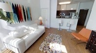 This single-floor space offers a clean, contemporary aesthetic defined by an open-plan layout that seamlessly connects the living area and kitchen. The living room is bright and airy, bathed in natural light that reflects off crisp white walls and light hardwood flooring. A white sofa anchors the seating area, complemented by a colorful patterned area rug and a warm-toned table lamp. A striking display of vertical surfboards in vivid hues adds a bold, coastal-inspired focal point to the space.

The kitchen is thoughtfully designed with rich dark wood cabinetry, stainless steel appliances including a refrigerator and microwave, and a peninsula-style breakfast bar fitted with sleek modern barstools. Pendant lighting hangs above the bar, providing both functional illumination and a stylish design detail. The kitchen's elevated countertop creates a natural visual division between the cooking and living zones while maintaining the open feel of the overall space.

The interior is characterized by high ceilings that amplify the sense of volume, white walls that enhance brightness, and a cohesive color palette of neutrals accented by pops of color through décor. The overall ambiance is modern, light-filled, and casually sophisticated with a relaxed coastal sensibility.
