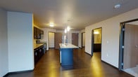 This single-floor space is thoughtfully designed with a modern aesthetic and an open-concept layout that maximizes functionality and flow. The kitchen serves as the centerpiece of the space, anchored by a sleek central island with a built-in sink and pendant lighting overhead, providing both a practical workspace and a visual focal point. Dark, rich cabinetry lines the kitchen walls, complemented by stone countertops and a full suite of stainless steel appliances, including a range, microwave, and refrigerator. A tiled backsplash adds a refined touch to the cooking area. The flooring throughout is a deep, dark hardwood that lends warmth and continuity to the open space, tying the kitchen and living areas together seamlessly. Recessed and flush-mount ceiling lighting ensures the space is well-lit throughout. Doorways lead to additional rooms, including what appears to be a bathroom visible from the main living area, featuring a vanity with mirror and warm lighting. The entry door is positioned at the far end of the open space, contributing to a practical and well-organized floor plan. Overall, the space presents a clean, contemporary character with cohesive design choices throughout.