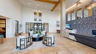 Located at 34 Ramsey Branch Rd in Freeport, Florida, this single-floor space presents a striking first impression through its dramatic, soaring ceilings accentuated by exposed natural wood beams and a sleek modern chandelier. The open living area is unified by large-format herringbone-patterned flooring in warm, light wood tones, creating a seamless and elegant foundation throughout the entire space.

Floor-to-ceiling black-framed glass partition walls and entry doors allow natural light to flow freely between zones while lending an industrial-chic aesthetic to the overall design. Oversized windows along the upper walls further flood the interior with daylight and offer views to the surrounding outdoor landscape.

The main lounge area features a carefully curated seating arrangement with cane-accented chairs and a bold circular coffee table, anchored by a layered area rug. A decorative accent wall clad in a graphic botanical wallcovering hosts arched mirrors and pendant lighting, adding depth and visual interest. Adjacent to this, a marble-topped reception counter provides a polished focal point.

A beverage station with dark wood cabinetry and a stainless steel under-counter refrigerator offers functional convenience, while an array of curated artwork displayed throughout adds cultural character. The space effortlessly balances refined design with a warm, welcoming ambiance.