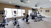This fitness center offers a comprehensive workout environment designed to accommodate a range of exercise needs. The space is outfitted with an assortment of cardio equipment, including an elliptical trainer and a recumbent exercise bike, providing options for low-impact cardiovascular workouts. A multi-station cable and weight machine anchors the strength training area, complemented by additional resistance equipment to support full-body conditioning routines.

Large windows line one wall of the room, flooding the space with natural light and offering views of the surrounding landscape, creating an open and energizing atmosphere. The neutral, warm-toned walls contribute to a welcoming ambiance, while the durable, textured flooring provides both safety and functionality underfoot.

Two ceiling fans are positioned throughout the room, ensuring consistent air circulation and a comfortable temperature during intense training sessions. Recessed lighting supplements the natural light, keeping the space well-lit at all times. A wall-mounted television is also present, allowing members to stay entertained during their workouts. The overall layout is thoughtfully organized to allow for ease of movement between equipment, making this fitness center a practical and inviting space for daily exercise.