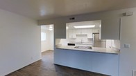 This single-story residence offers a clean, modern aesthetic throughout its thoughtfully designed layout. The heart of the home is an open-concept kitchen, highlighted by a skylight that floods the space with natural light. White cabinetry lines the walls, providing ample storage, while light-toned countertops offer generous workspace. Stainless steel appliances, including a refrigerator and cooktop, lend a contemporary touch, complemented by a stainless steel sink set beneath the cabinetry. A peninsula-style counter with a sleek blue-gray base extends into the living area, creating an ideal space for casual dining or entertaining while maintaining an open, connected feel. Rich, dark hardwood flooring anchors the main living and kitchen areas, adding warmth and contrast to the light walls and cabinetry. A wide pass-through opening connects the kitchen to an adjacent room, which features lighter flooring and a window that brings in additional natural light. The overall ambiance is bright, neutral, and move-in ready, with a practical and streamlined design that balances functionality with a contemporary residential style.