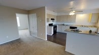 This single-floor residential unit offers a practical and well-organized open-concept layout designed for comfortable everyday living. The living area is finished with wall-to-wall carpet, creating a warm and welcoming atmosphere, while the adjacent kitchen features dark hardwood-style flooring that provides a clean visual contrast.

The kitchen is equipped with white cabinetry offering ample storage, a black gas range with overhead microwave ventilation, a dishwasher, and a peninsula counter with granite-style countertops — ideal for meal preparation and casual dining. A ceiling fan positioned above the kitchen and living area transition ensures consistent airflow throughout the shared space.

Neutral beige walls and a textured ceiling run throughout the unit, lending a cohesive and understated aesthetic. The living area opens to a bedroom, which benefits from natural light via a window, and connects to a full bathroom also featuring natural light. A closet with bi-fold doors is conveniently accessible from the main living space.

The floor plan is thoughtfully arranged to maximize functionality, with clear sight lines between the kitchen, living, and sleeping areas. The unit's design provides a straightforward, low-maintenance living environment suited for a range of occupants.
