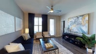 This 1-bedroom, 1-bathroom residence is designed with modern sensibilities and functional living in mind. The living area serves as the centerpiece of the home, featuring rich hardwood-style flooring that runs throughout and neutral-toned walls that create a warm, inviting atmosphere. A ceiling fan with a sleek fixture provides both comfort and a contemporary aesthetic touch overhead. Large windows dressed with floor-to-ceiling dark curtains invite an abundance of natural light while offering flexible privacy options. A glass-paneled door leads to an outdoor access point, further enhancing the sense of openness. The living space is generously proportioned, offering ample room for a full sofa arrangement, accent seating, and a media console, making it well-suited for both relaxing and entertaining. The overall ambiance is polished and modern, with clean lines and a neutral palette that lends itself easily to personalization. The single-floor layout ensures seamless flow between spaces, making everyday living both convenient and comfortable. This residence combines practical design with stylish details for an elevated everyday living experience.