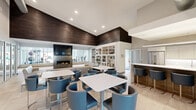 Located at 2801 Stonebridge Blvd in New Castle, Delaware, this thoughtfully designed single-floor community space offers a sophisticated and functional environment for residents. The open-concept layout seamlessly integrates a spacious lounge and a fully equipped kitchen, creating a welcoming communal hub.

The living area is a standout, anchored by a sleek linear fireplace with a wall-mounted television above, flanked by expansive windows that overlook an adjacent fitness center. Low-profile, neutral-toned sofas are arranged to encourage conversation, while illuminated built-in window benches add ambient lighting along the perimeter. A dramatic floor-to-ceiling white bookshelf wall lined with curated décor and books adds warmth and character, complemented by a rich dark wood plank ceiling overhead.

The kitchen is outfitted with stainless steel appliances, including a full-size French door refrigerator, clean white upper cabinetry, warm wood lower cabinetry with brass hardware, and an extended white quartz countertop that doubles as a serving bar. Pendant lighting and large-format tile flooring throughout reinforce the modern aesthetic.

Glass-walled private office or meeting rooms line one side of the space, offering visibility while maintaining separation. The overall ambiance is bright, clean, and contemporary — designed to foster both relaxation and productivity.