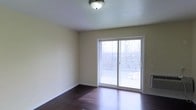 This single-floor residence offers a clean, functional layout with a warm and inviting aesthetic. The living area is finished with rich, dark hardwood flooring that anchors the space and provides a striking contrast against the soft, neutral cream-toned walls. Natural light pours into the room through a large sliding glass door, which serves as both a visual focal point and a direct access point to a private balcony. The balcony, enclosed by a wrought-iron railing, overlooks a scenic landscape of trees and rolling terrain, providing a tranquil outdoor retreat. The ceiling is smooth and white, fitted with a classic flush-mount light fixture that complements the understated, practical design of the space. A wall-mounted heating unit is also present, adding to the room's functional features. The overall ambiance is bright, open, and move-in ready, with a straightforward design that allows for flexible furnishing and personalization. The combination of hardwood floors, abundant natural light, and outdoor access makes this a comfortable and well-appointed living space.