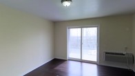 This single-floor residence offers a clean, functional layout with a warm and inviting aesthetic. The living area is finished with rich, dark hardwood flooring that anchors the space and provides a striking contrast against the soft, neutral cream-toned walls. Natural light pours into the room through a large sliding glass door, which serves as both a visual focal point and a direct access point to a private balcony. The balcony, enclosed by a wrought-iron railing, overlooks a scenic landscape of trees and rolling terrain, providing a tranquil outdoor retreat. The ceiling is smooth and white, fitted with a classic flush-mount light fixture that complements the understated, practical design of the space. A wall-mounted heating unit is also present, adding to the room's functional features. The overall ambiance is bright, open, and move-in ready, with a straightforward design that allows for flexible furnishing and personalization. The combination of hardwood floors, abundant natural light, and outdoor access makes this a comfortable and well-appointed living space.