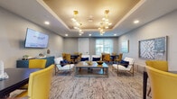 This thoughtfully designed communal space offers a welcoming and versatile environment suited for dining, socializing, and relaxation. The layout flows seamlessly across multiple distinct zones, beginning with a grand, double-height dining hall bathed in natural light from tall windows. Elegant chandeliers and sputnik-style pendant lights hang from the soaring ceilings, casting a warm glow over rows of dark wood dining tables paired with cream upholstered chairs and plush patterned area rugs.

Transitioning into the lounge area, a tray ceiling with a gold-painted inset adds architectural depth, accented by striking geometric brass pendant fixtures. Seating here includes a mix of bold mustard yellow accent chairs, crisp white armchairs with navy cushions, and intimate dining sets with amber leather chairs — all centered around low coffee tables and complemented by a teal credenza beneath a wall-mounted television.

A separate, more intimate dining nook features blue upholstered chairs around light wood tables, adorned with framed abstract artwork in cool coastal tones. A dedicated game and activity area with a pegboard display and bookshelf adds a recreational dimension to the space. Throughout, light hardwood flooring and neutral carpeting tie each zone together, while curated artwork and lush greenery contribute to the overall refined, contemporary ambiance.