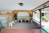 This common area lobby presents a distinctive Mediterranean-inspired aesthetic that immediately captures attention. The entry features large floor-to-ceiling glass doors framed in dark metal, offering views of lush outdoor greenery, palm trees, and the surrounding courtyard beyond. Pendant lantern-style light fixtures flank the entrance, enhancing the welcoming character of the space.

A prominent staircase with teal-painted metal railings, accented with decorative scrollwork, connects the levels and serves as a central architectural feature. The stairs are finished with terrazzo-style treads, complementing the warm terracotta tile flooring found throughout the lower level.

The walls are adorned with vibrant hand-painted murals depicting scenic landscapes, archways, trees, and garden motifs in warm earth tones, adding a lively artistic dimension to the space. These murals extend across multiple curved and flat wall surfaces, creating a cohesive and immersive visual environment.

The upper landing area features a carpeted lounge space with stone benches and potted tropical plants arranged beneath the mural backdrop, offering a relaxed seating area bathed in natural light from the adjacent glass entry. Textured ceilings and wall-mounted lighting fixtures complete the well-maintained, character-rich environment throughout.
