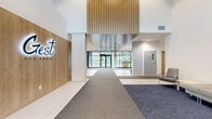 Located at 3551 Boulevard Gouin Est in Montréal, this lobby and lounge space offers a refined and contemporary first impression. The main entry corridor features large-format white tile flooring, layered carpet runners, and soaring double-height ceilings framed by floor-to-ceiling glazed walls that flood the space with natural light and provide clear views of the surrounding greenery. A striking slatted wood ceiling detail and a branded feature wall add architectural character to the arrival experience.

Transitioning from the entry hallway, black-framed glass partitions elegantly define the lounge area while maintaining visual openness throughout. The lounge is designed for comfort and social interaction, with warm hardwood flooring, circular pendant lighting, and a mix of seating arrangements including plush sofas, colorful accent chairs, and intimate groupings around area rugs. Built-in shelving with deep teal cabinetry, a wall-mounted display, and a linear fireplace contribute to the space's inviting ambiance.

Additional areas extend the lounge's functionality, offering quieter zones with individual workstations and access to outdoor terraces through full-height glazed doors. Lush trailing greenery suspended from black metal frames adds a biophilic touch, softening the modern aesthetic and enhancing the overall warmth of this thoughtfully designed communal space.