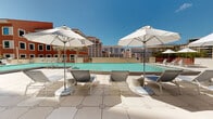 This sophisticated outdoor amenity level is designed for resort-style urban living, offering a thoughtfully curated collection of spaces set against a stunning city skyline backdrop. At its centerpiece sits a generously sized swimming pool with crystal-clear turquoise water, flanked by sun loungers and shaded by classic white market umbrellas — an ideal setting for relaxation beneath open skies. Adjacent to the pool, a dedicated outdoor kitchen features a built-in stainless steel grill and ample counter space, complemented by a dining area furnished with modern tables and chairs, perfect for al fresco entertaining. A separate garden lounge area provides a more intimate retreat, featuring built-in wood-accented bench seating, lush tropical landscaping with soaring palm and cypress trees, and warm string lighting overhead that creates an inviting ambiance for evening gatherings. A fire feature anchors this seating area, adding a distinctive focal point. Throughout the deck, large-format light stone pavers and crisp white railings maintain a cohesive, elegant aesthetic. The surrounding multi-story building frames the space with balconied facades and tropical potted plantings, while panoramic views of the urban skyline complete this premier outdoor destination.