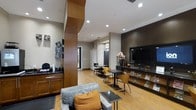The Media Cafe is a thoughtfully designed, single-floor communal space that blends comfort and functionality in a contemporary setting. The layout is open and inviting, with warm bamboo-style hardwood flooring running throughout that contrasts elegantly against the darker accent walls and cabinetry.

A comfortable lounge area anchors one side of the space, featuring a plush gray sofa accented with vibrant orange throw pillows and paired with a sleek glass side table — ideal for casual relaxation. Directly across, a large flat-screen television is mounted on a dark feature wall, surrounded by a media console with built-in shelving that displays reading materials and decorative accents.

The café counter area is equipped with a dark granite countertop, wood cabinetry, and what appears to be a beverage station, providing a convenient self-serve setup for residents or guests. Adjacent to the lounge, a dining area features modern black tables paired with contemporary amber-toned chairs, offering a practical space for casual meals or informal gatherings.

Recessed ceiling lighting throughout ensures the space is well-lit, while the warm wood tones, neutral walls, and cohesive furnishings create an atmosphere that is both polished and approachable. The Media Cafe is well-suited for unwinding, socializing, or staying informed and entertained.