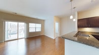 This single-floor space offers a clean, modern layout designed for comfortable everyday living. The open-concept floor plan seamlessly connects the kitchen and living area, creating a sense of spaciousness and ease of flow throughout. Rich hardwood flooring extends across the main living space, adding warmth and a polished finish to the interior.

The kitchen is appointed with dark granite countertops and features a raised breakfast bar, ideal for casual dining or entertaining. Stylish pendant lighting hangs above the counter, providing both functional illumination and a contemporary design accent.

The living area is generously sized, bathed in natural light streaming through large windows and a sliding glass door that opens to an outdoor balcony area. The neutral wall tones and white trim throughout the space create a bright, airy ambiance that complements the overall modern aesthetic.

A hallway visible from the main living area leads to additional rooms, suggesting a well-organized and thoughtfully designed layout. The space maintains a cohesive, move-in-ready feel with clean lines, neutral finishes, and an inviting atmosphere suited to a variety of lifestyles.
