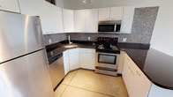 This two-floor loft-style space combines industrial character with modern finishes to create a distinctive and functional living environment. The main level showcases an open-concept kitchen outfitted with stainless steel appliances, including a range, microwave, refrigerator, and dishwasher. Rich dark granite countertops contrast elegantly with crisp white cabinetry, while a mosaic tile backsplash adds texture and visual interest. Large-format tile flooring runs throughout the main level, complementing the urban aesthetic. Exposed industrial ductwork runs overhead, and track lighting illuminates the space, reinforcing the loft's contemporary character. Soaring ceilings on the main level create an airy, expansive atmosphere. A staircase leads to the upper level, where sloped, wood-paneled ceilings lend an intimate architectural quality. The upper loft area features warm carpet flooring and offers a versatile space suitable for a sleeping area or lounge. An additional carpeted room on the lower level provides flexible use, complete with closet access. The full bathroom is finished with terracotta-toned tile flooring, a white vanity, and a full tub-shower combination, offering a clean and functional design. Throughout both levels, the interplay of industrial details and refined finishes gives this space a bold, urban personality.