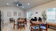 This single-floor communal space is thoughtfully designed to serve as both a relaxed lounge and a casual café-style gathering area. The interior features vaulted ceilings that lend an airy, open feel throughout, complemented by light-toned hardwood flooring that runs seamlessly across the entire space. Recessed lighting and a modern ceiling fan with sleek black blades provide both illumination and comfort.

The main area is furnished with multiple round dining tables paired with mid-century modern wood and upholstered chairs, creating an inviting atmosphere for socializing or casual meetings. A dedicated coffee station, marked by a decorative wall-mounted "Coffee" sign, is anchored by a dark credenza housing coffee makers and supplies, accompanied by a utility wet bar area with dark cabinetry, white countertops, and a stainless steel sink for added convenience.

A lounge nook provides a quieter retreat, featuring leather armchairs, a corner fireplace, and abundant windows that invite natural light and views of surrounding greenery. Tasteful framed artwork, decorative potted plants, and carefully placed accent lamps complete the warm, contemporary ambiance throughout the space.