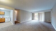 This single-floor unit offers a practical and well-organized layout across several distinct living spaces. The main living area is generously proportioned, featuring neutral gray walls, plush wall-to-wall carpeting, and a large sliding glass door fitted with vertical blinds that allows natural light to fill the room while providing access to an outdoor view of greenery. A secondary carpeted room, visible from the living area, offers additional flexible living space with its own entry door.

The kitchen is efficiently designed with warm oak cabinetry extending to the upper and lower walls, a white gas range, a double-basin sink, and light tile flooring that contrasts with the carpeted areas throughout the rest of the unit. Ample counter and storage space make the kitchen highly functional.

The bedrooms are carpeted and feature neutral-toned walls, standard windows with blinds that bring in natural light, and ceiling-mounted light fixtures. One bedroom includes a built-in closet with shelving, offering practical storage. The full bathroom is clean and straightforward, equipped with a shower-tub combination, a toilet, and a vanity with storage below. White walls and tile flooring give the bathroom a bright, utilitarian feel throughout.