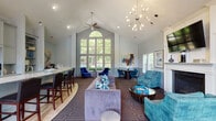 Located at 100 Shore Lake Dr in Greensboro, NC, this single-story clubhouse presents a thoughtfully designed gathering space with a refined, contemporary aesthetic. The main lounge area commands attention with dramatically vaulted ceilings that rise to a striking peak, anchored by a statement sputnik chandelier and complemented by a ceiling fan. A towering arched window wall floods the space with natural light and frames lush outdoor greenery, creating a seamless connection with the surrounding landscape.

The interior is finished in soft blue-grey tones with wainscoting panels lining the lower walls throughout, lending a polished, traditional character. The patterned carpet in deep brown with geometric line motifs flows across the main areas, contrasting with warm hardwood flooring near the bar. A curved bar counter with a marble-look surface and built-in grey cabinetry with open display shelving serves as a functional centerpiece.

Distinct seating zones are thoughtfully arranged, including a lounge area with plush teal and lavender upholstered furniture, a café-style dining nook, and a quieter sitting room featuring a fireplace with a mounted television, a round table, and direct outdoor access. Natural light pours in from multiple windows throughout, while curated artwork adds vibrant color accents across the space.