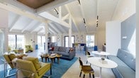 This single-story space makes a striking architectural statement with its soaring vaulted ceiling defined by exposed, white-painted structural beams and trusses. Track lighting runs along the ceiling framework, providing both ambient and accent illumination throughout the open-plan interior. The white shiplap ceiling and light-toned hexagonal tile flooring work together to create a bright, airy atmosphere.

The open living area is anchored by a bold blue area rug, upon which sits a contemporary gray sofa accented with patterned and colored throw pillows. Complementing this arrangement is a pair of chartreuse yellow accent chairs with chrome frames, reflecting a confident mid-century modern design sensibility. A cluster of organic-shaped wooden coffee tables adds warmth to the seating area.

A round white dining table accompanied by molded wooden chairs occupies one corner, while a bar-height table with stools defines a casual dining or workspace nearby. Decorative metallic sphere wall accents add a playful, artistic touch. Large windows and glass doors on multiple walls flood the interior with natural light and offer views of the surrounding trees, enhancing the connection to the outdoors.